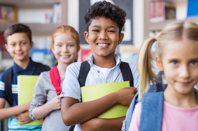 Elementary students holding books and wearing backpacks in front of bookshelves.